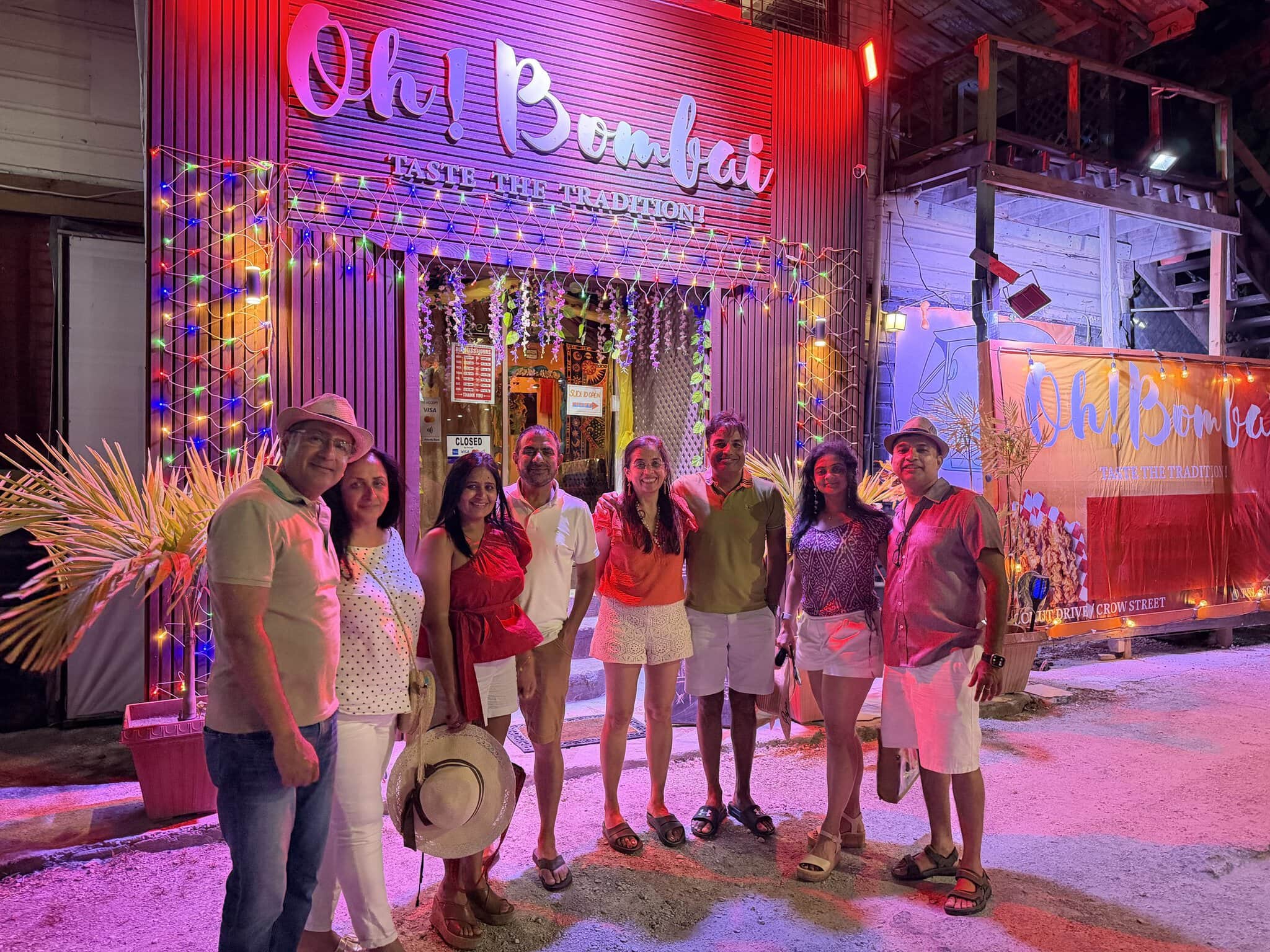 Group of eight friends outside Oh! Bombai Indian restaurant at night in San Pedro, Belize, with the lit neon sign reading Oh! Bombai - Taste the Tradition