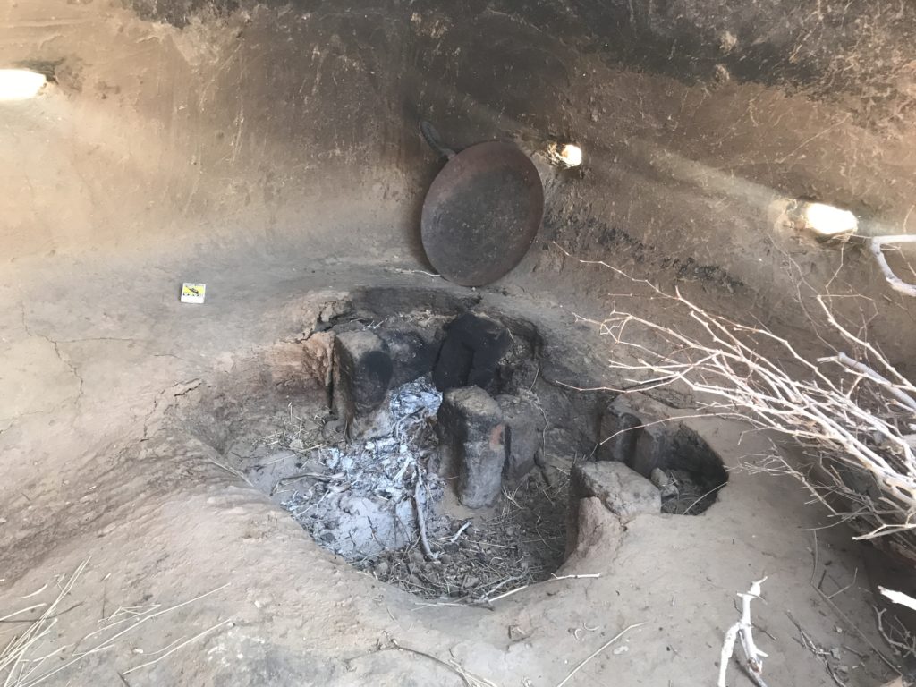 A wood fire stove, tava and firewood in an otherwise empty kitchen. 