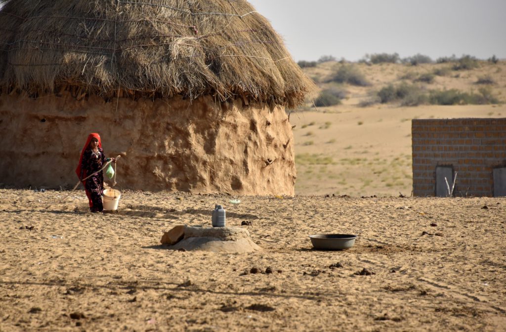 A girl fetching water from a kund (well) near her large hut shaped house. 