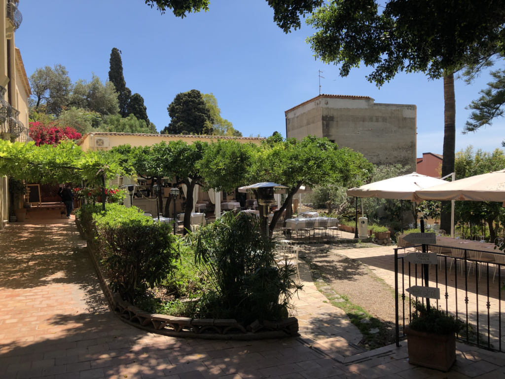 A traditional outdoor garden setting with meeting rooms along the left covered walkway and casually spread out tables on multiple levels. - at a restaurant in Sicily.