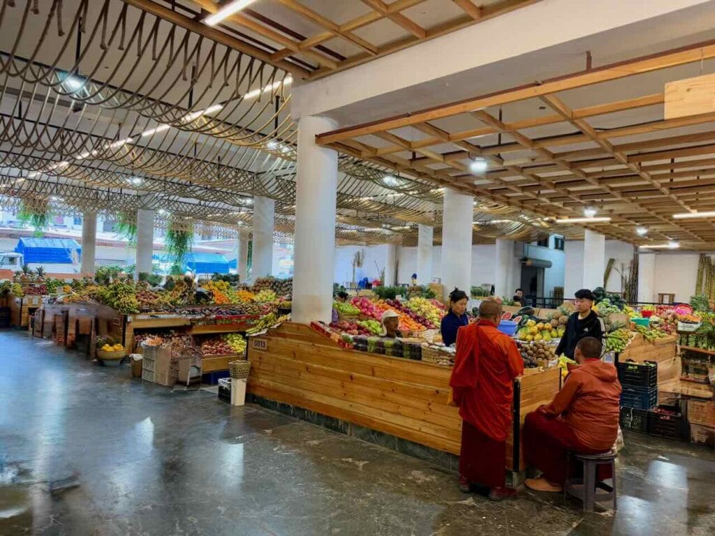 Food market in Thimpu, Bhutan, with rows of stalls piled with colorful vegetables. A couple of Buddhist monks are shopping at a stall
