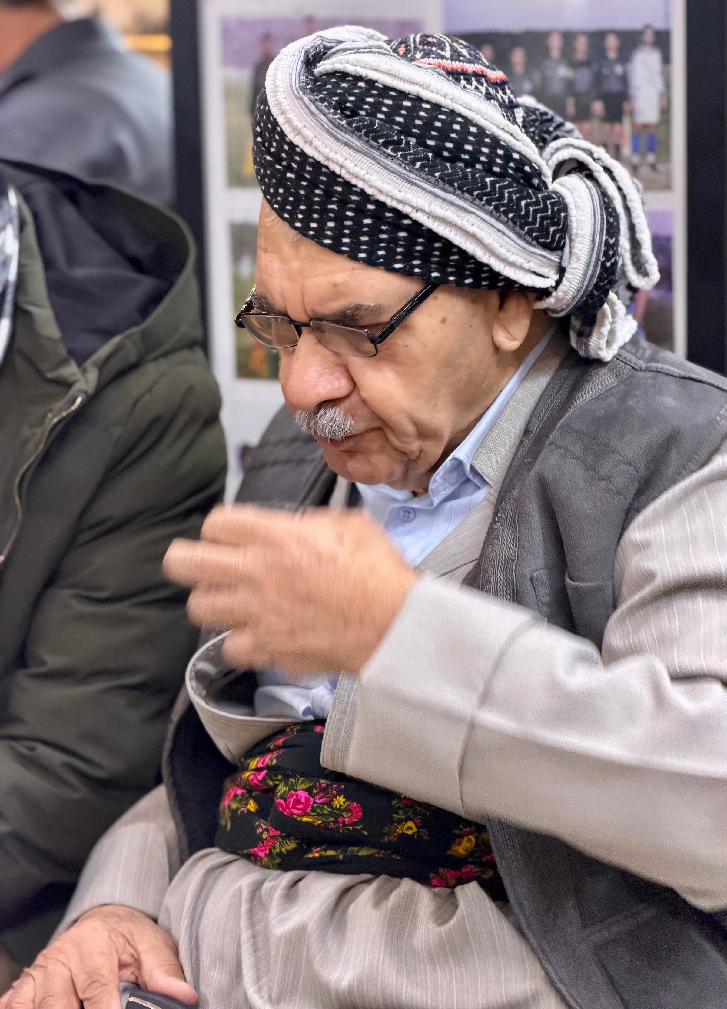 An elderly Kurdish man in traditional dress with keffiyeh and embroidered sash gesturing in conversation at the Erbil bazaar, Iraqi Kurdistan