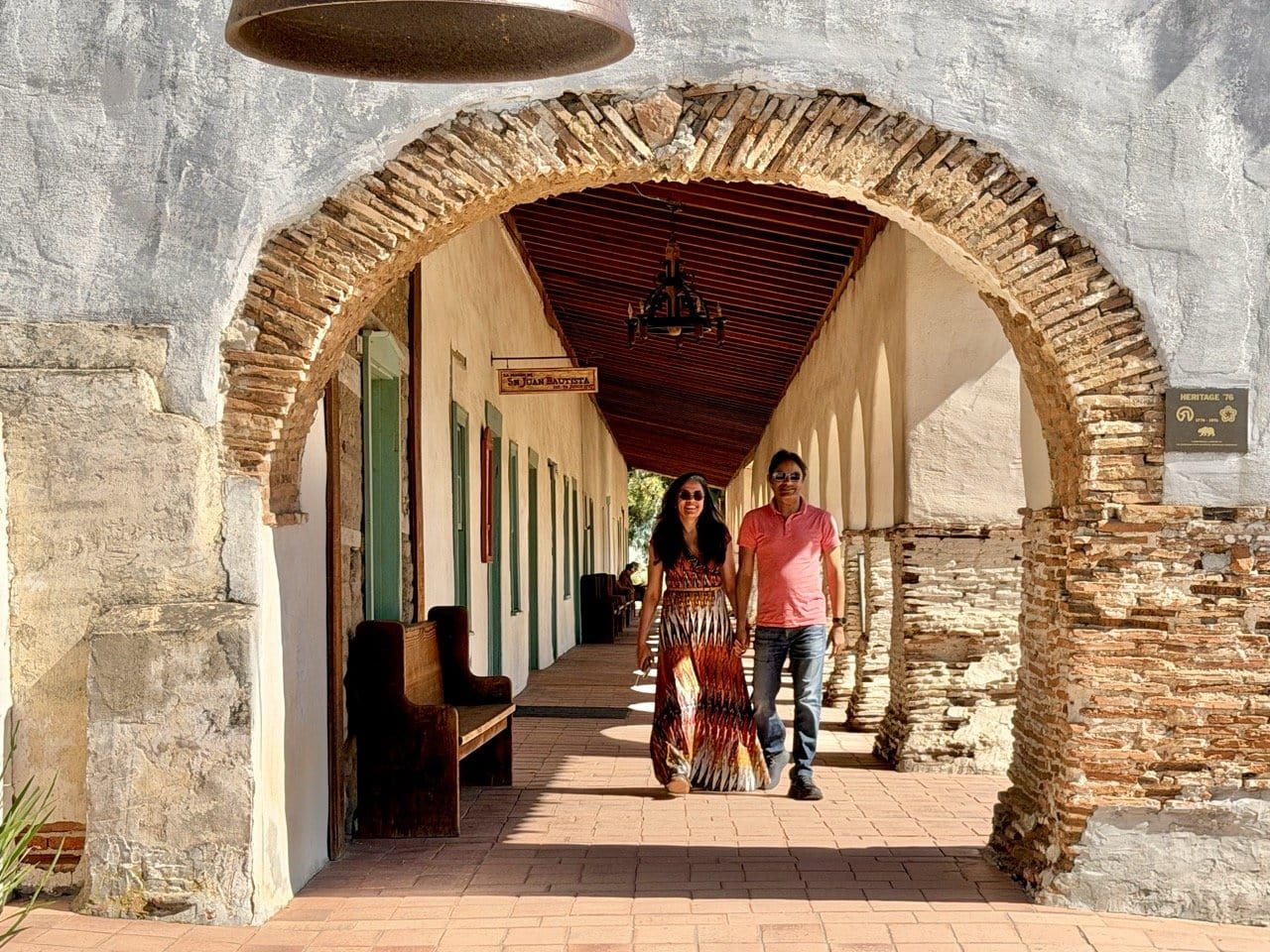 Nirmal and Jyoti walking through the stone arch colonnade at Mission San Juan Bautista with brick pillars and wooden ceiling receding into the distance