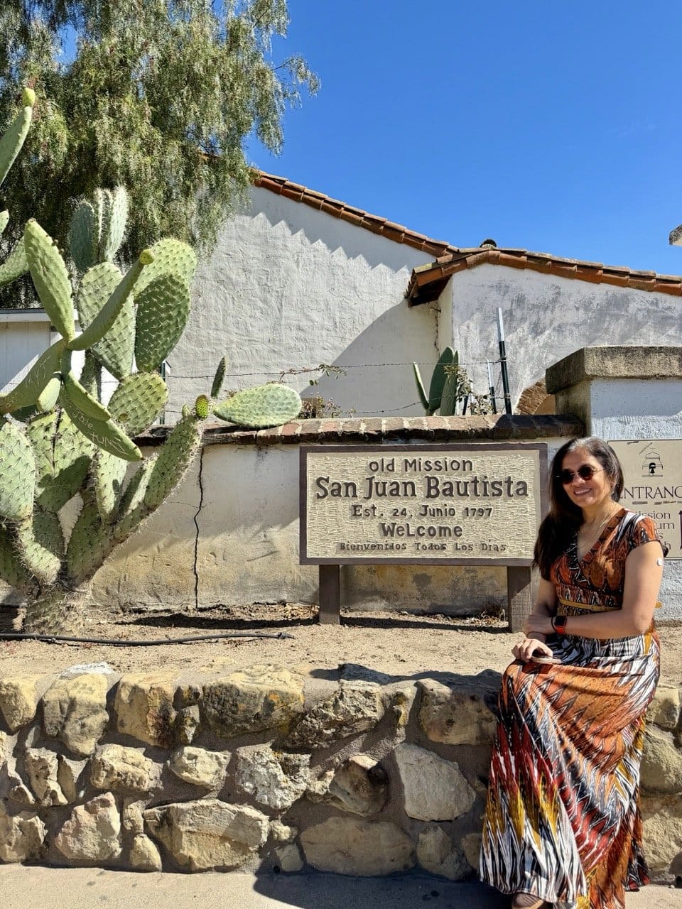 Jyoti sitting beside the Old Mission San Juan Bautista entrance sign with a large prickly pear cactus and the whitewashed mission wall behind