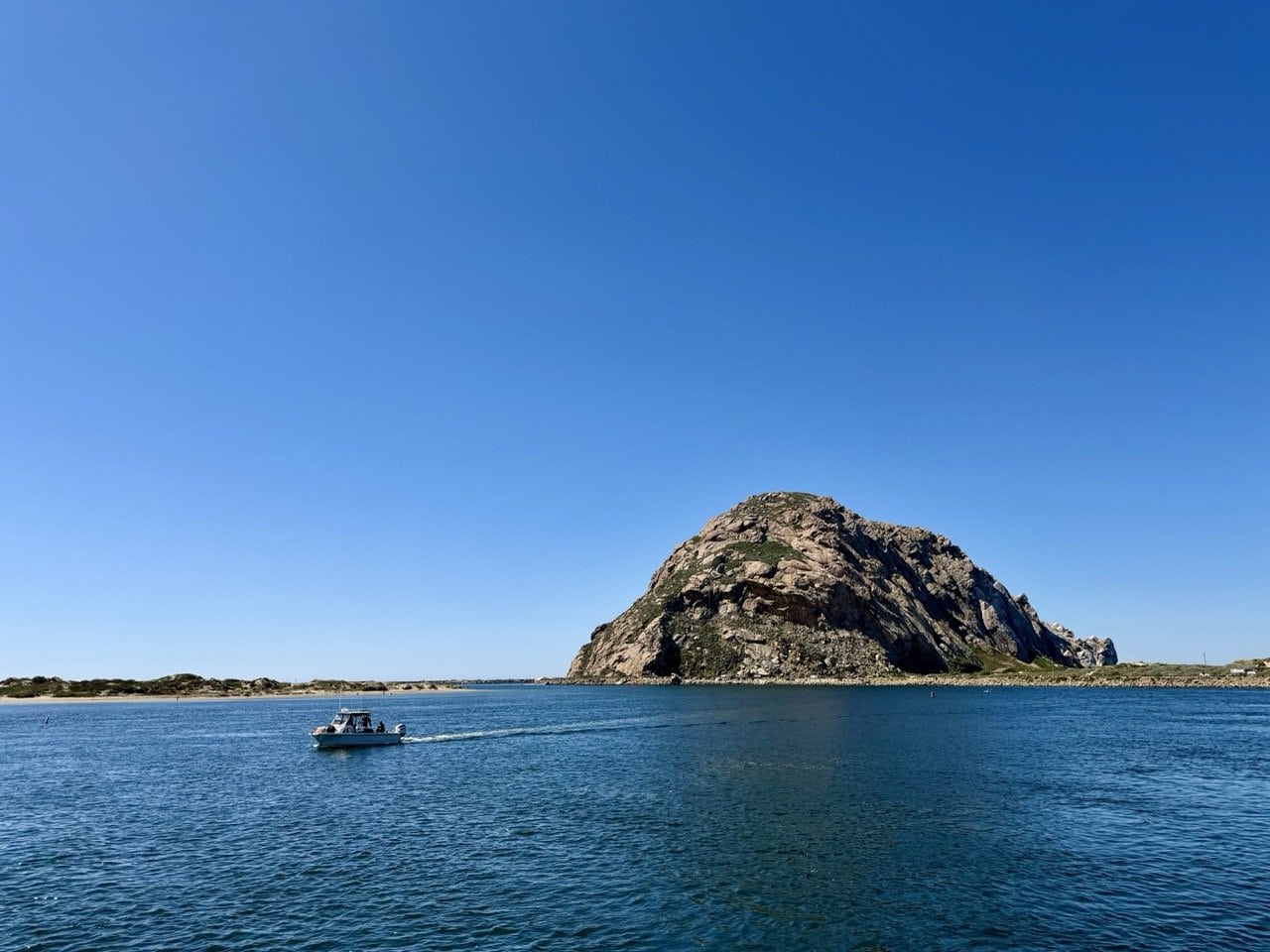 Morro Rock rising from the calm blue waters of Morro Bay with a small boat crossing in the foreground and a deep blue sky above