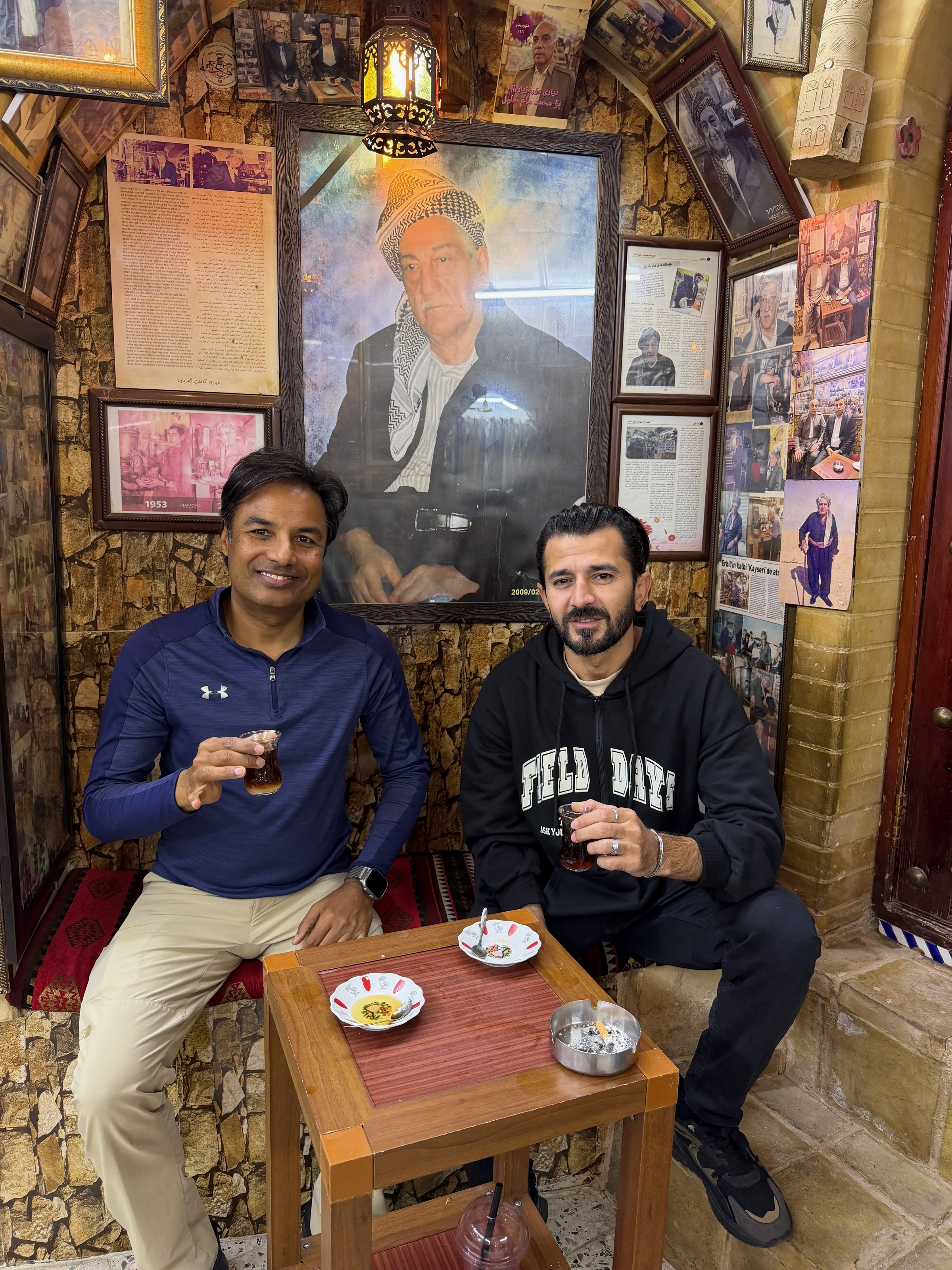 Nirmal and his Kurdish guide sharing tea at a historic tea house in the Erbil Citadel bazaar, walls and ceiling covered in framed photographs and Kurdish portraits, Iraqi Kurdistan