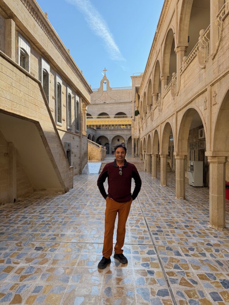 Nirmal standing in the tiled courtyard of Mar Mattai Monastery with arched colonnades and a church cross visible behind him, northern Iraq