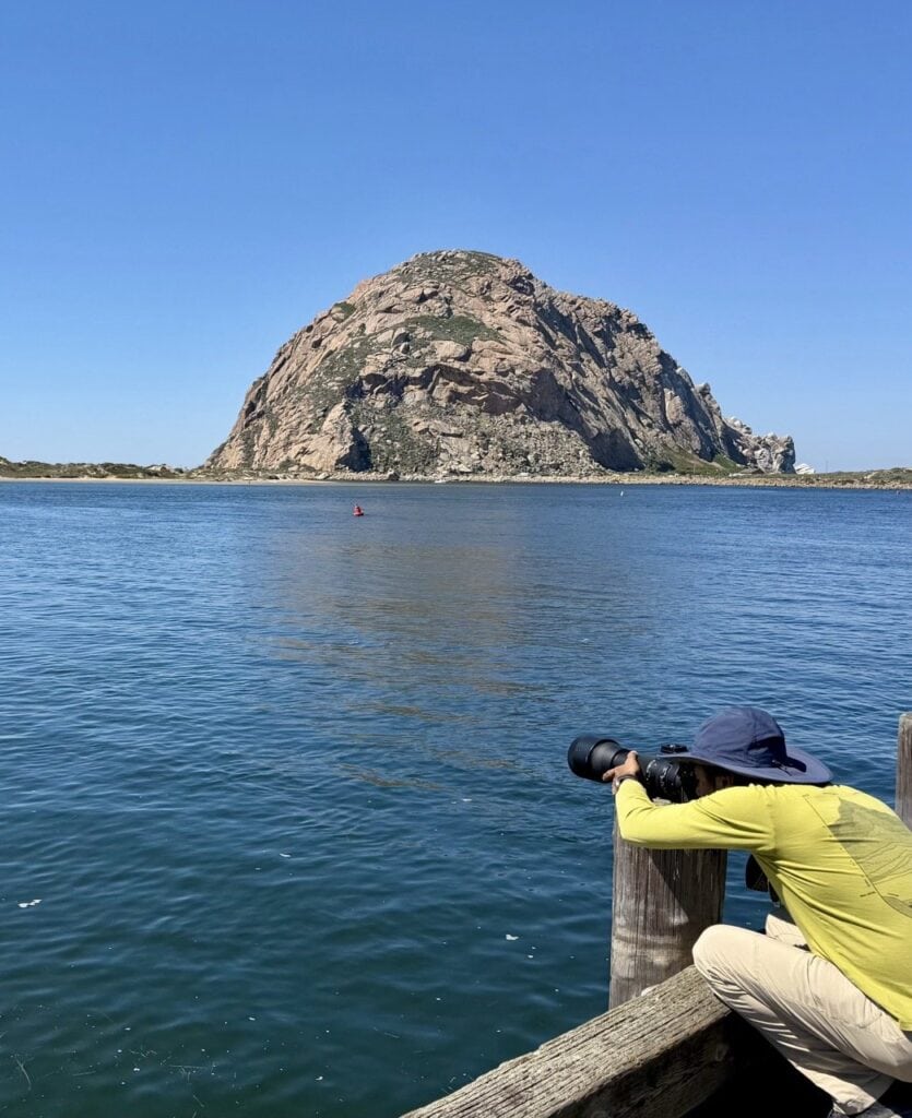 Nirmal crouched at the dock railing with his long telephoto lens trained on the water searching for sea otters with Morro Rock rising dramatically behind him