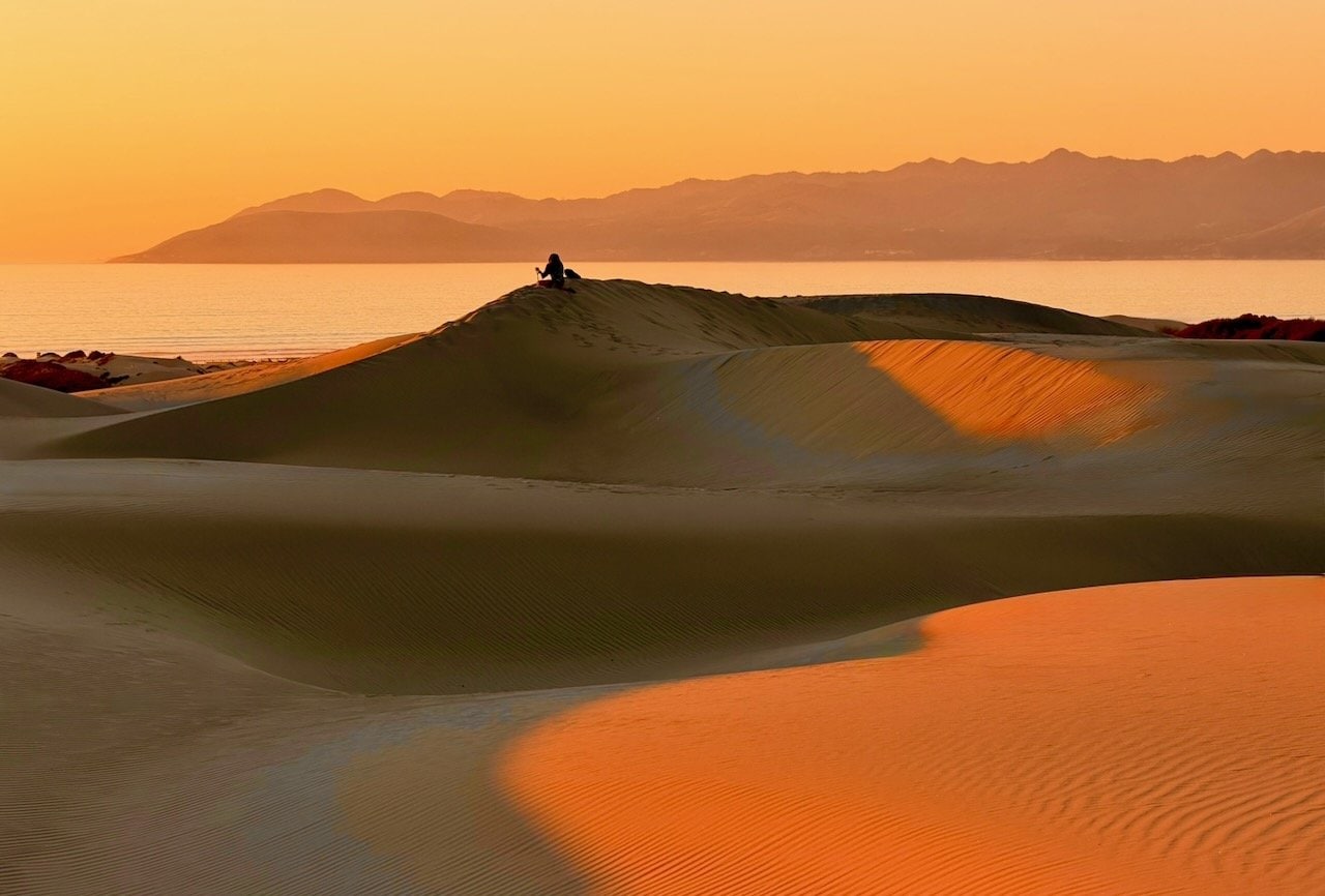 Two bare footprints pressed into orange-red sand at Oceano Dunes with the setting sun visible over the dunes in the background