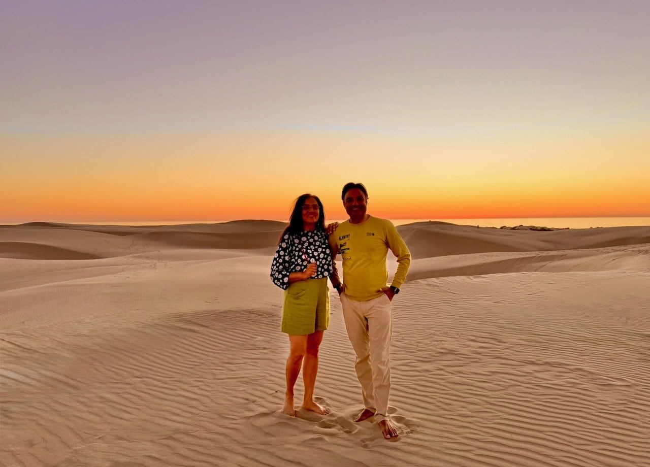 Nirmal and Jyoti standing barefoot on the sand dunes at Oceano at dusk with a pastel orange and purple sky and ocean on the horizon behind them