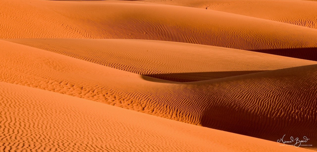 Wide view of Oceano sand dunes glowing deep orange at sunset with parallel ridges of rippled sand receding into the distance and mountains behind
