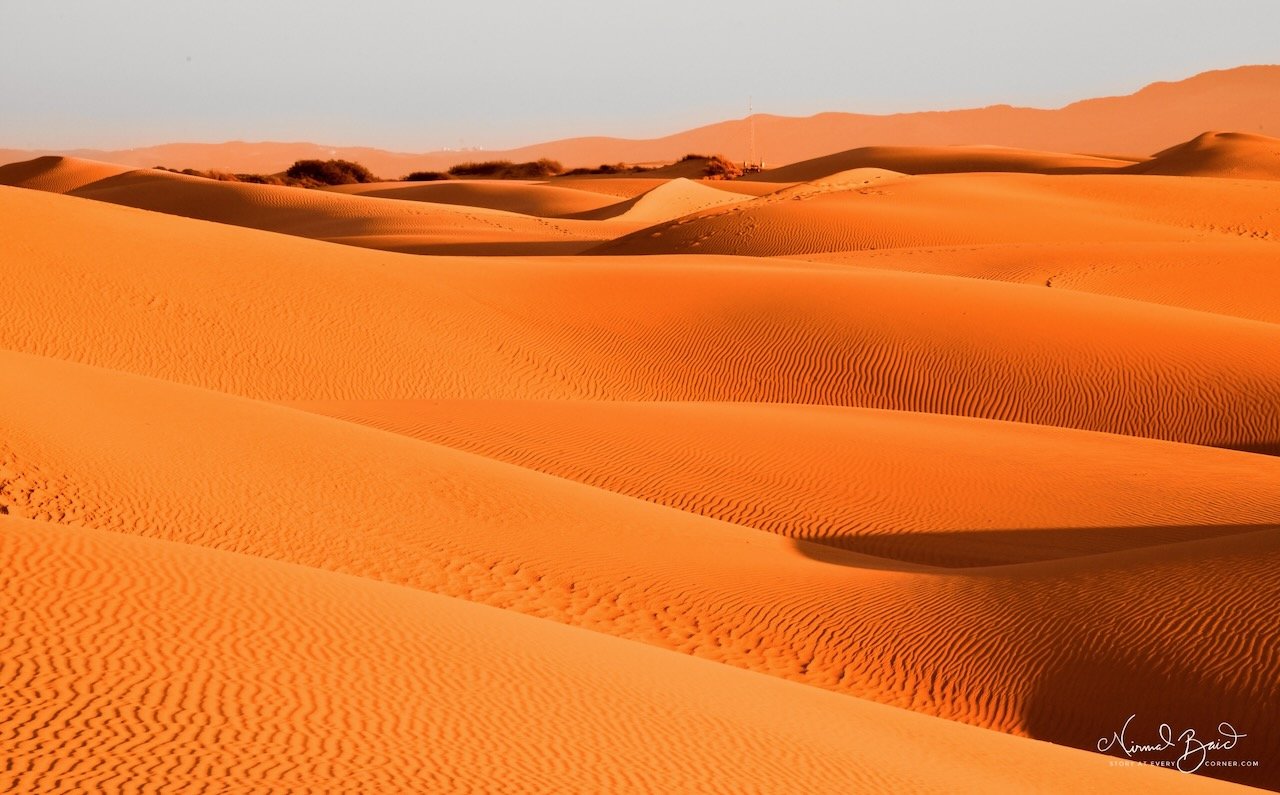 Panoramic view of multiple layered sand dune ridges at Oceano glowing orange-gold in warm sunset light with hazy mountains in the background