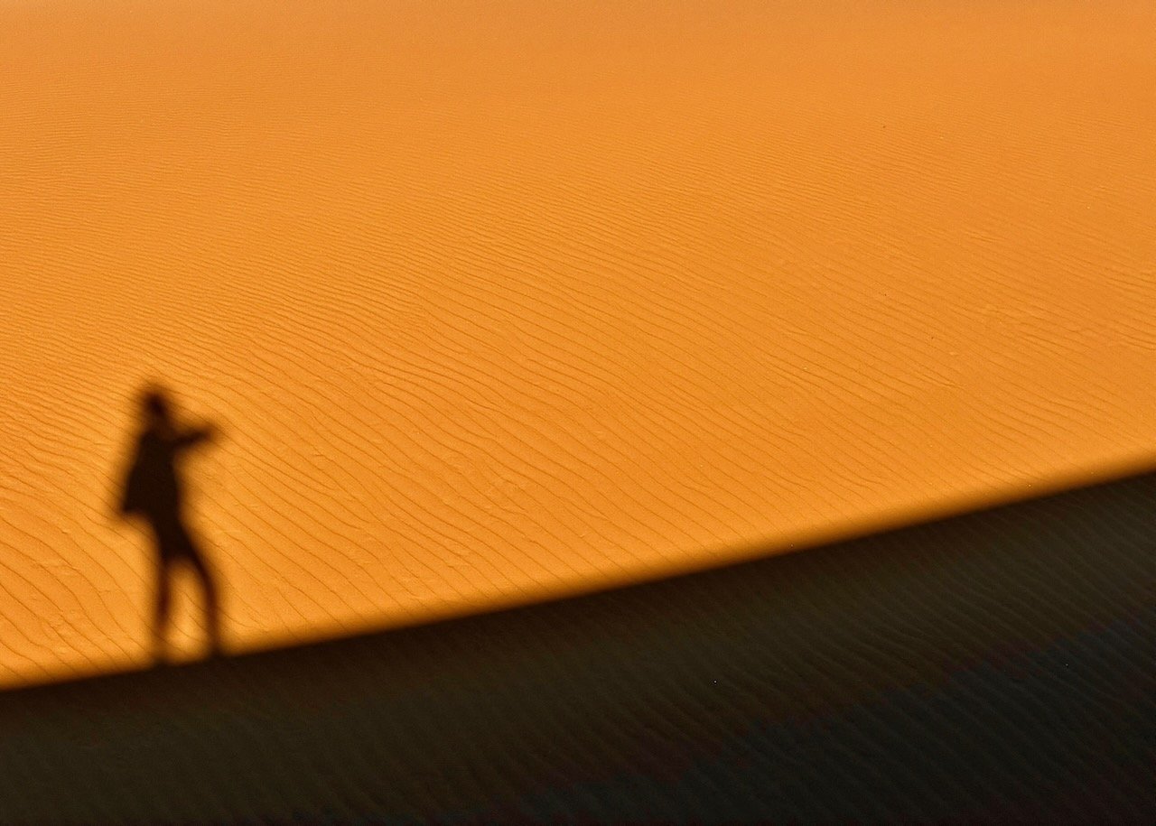 Shadow of a person standing on a sand dune ridge cast across the smooth orange surface of Oceano Dunes at sunset