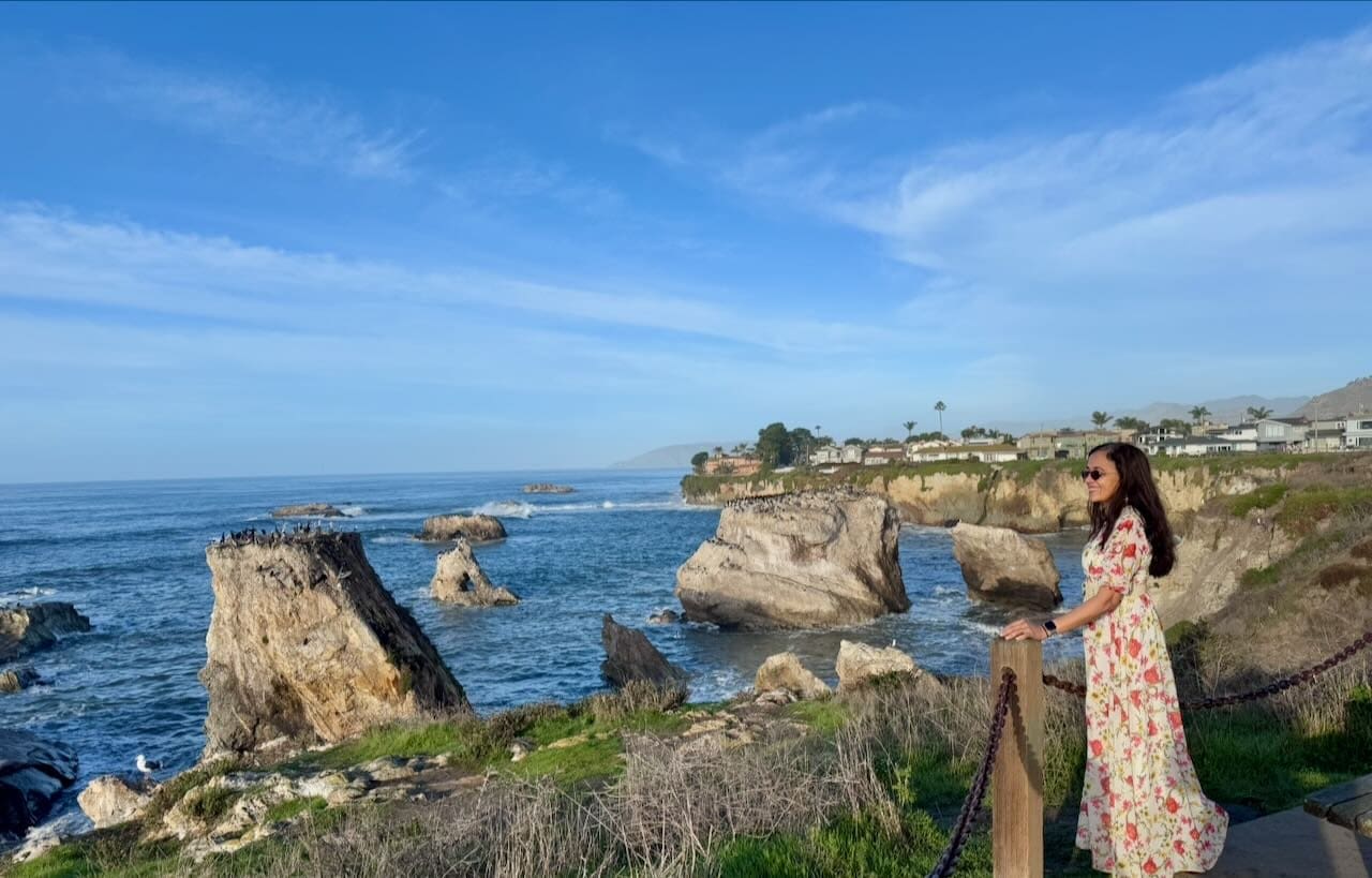 Jyoti standing at the Dinosaur Caves cliff viewpoint in Pismo Beach with dramatic sea stacks covered in seabirds and the Pacific Ocean behind her