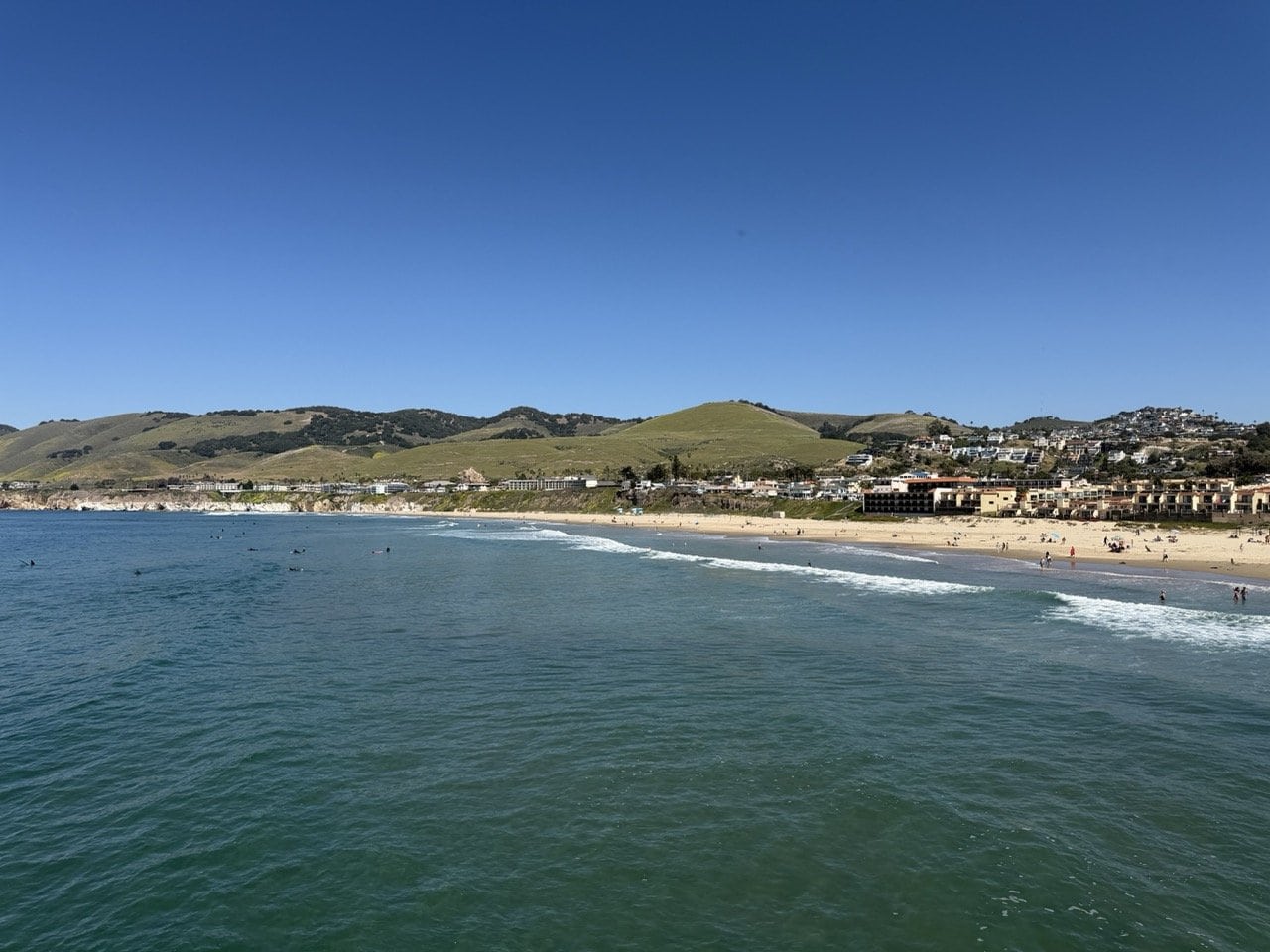 View of Pismo Beach from the end of the pier showing surfers in the water the sandy beach the town and green hills rising behind on a clear blue day