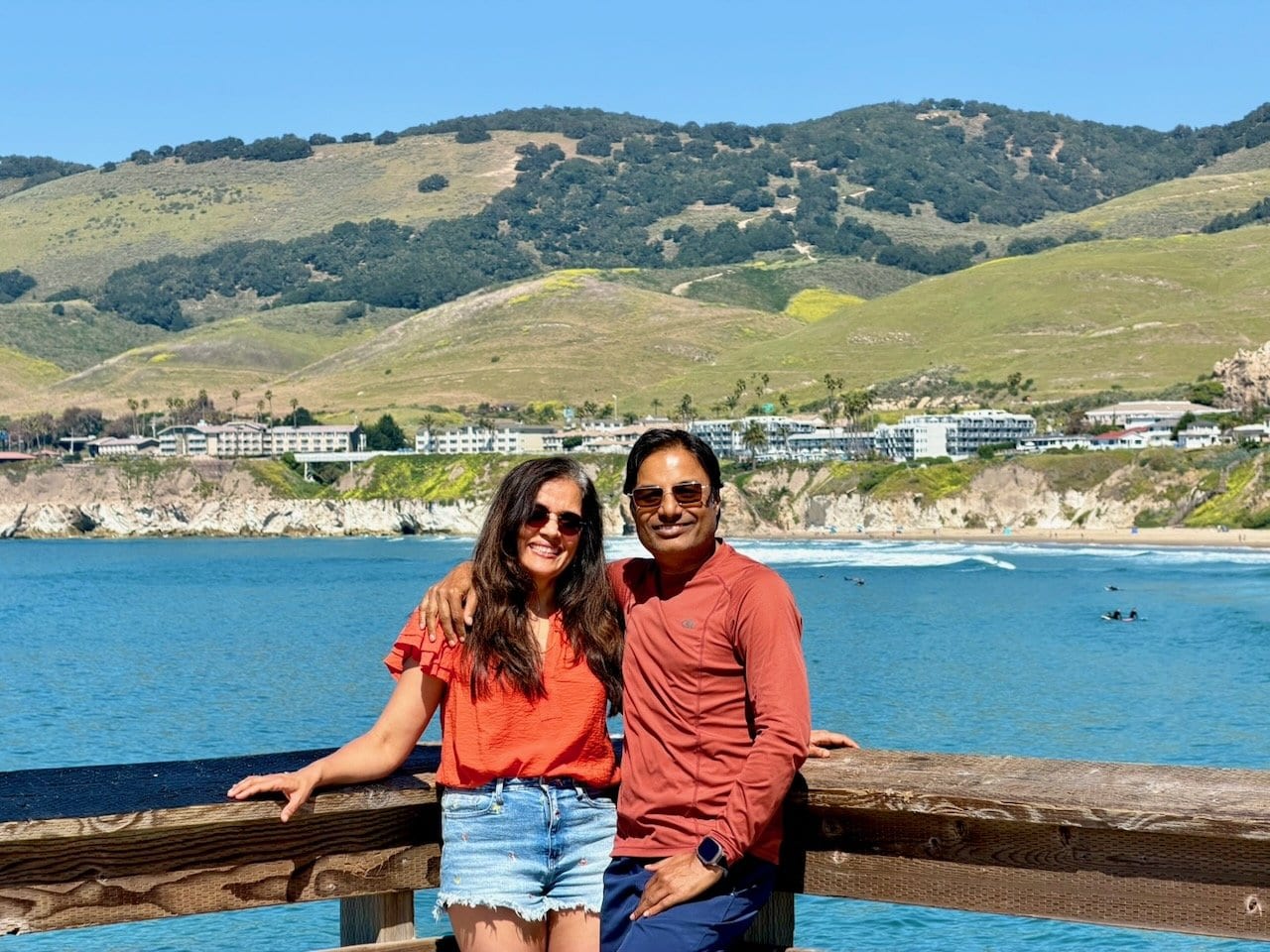 Nirmal and Jyoti smiling together on Pismo Beach pier with the blue bay surfers and green spring hills behind them