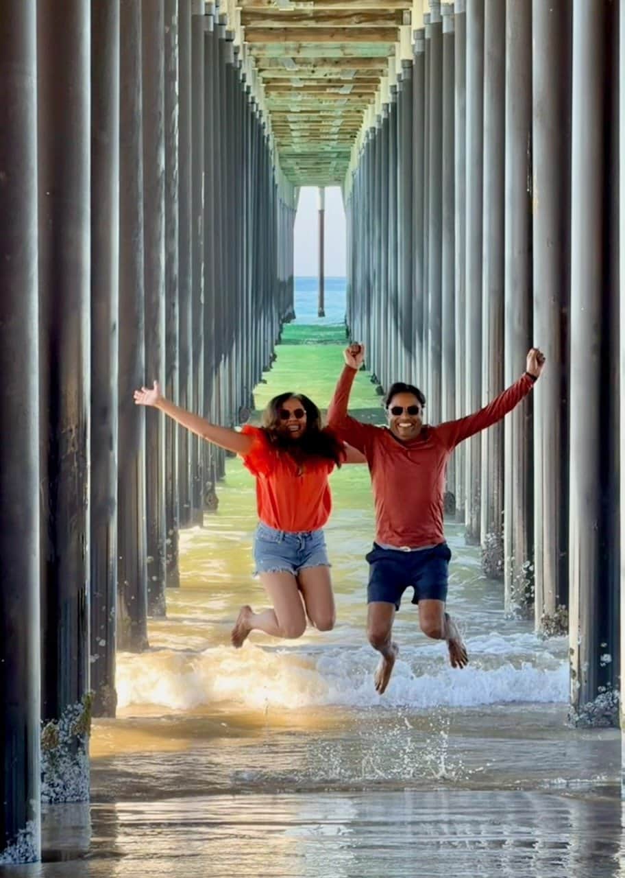 Nirmal and Jyoti walking hand in hand into the perspective tunnel beneath Pismo Beach pier with waves at their feet and ocean light glowing at the far end