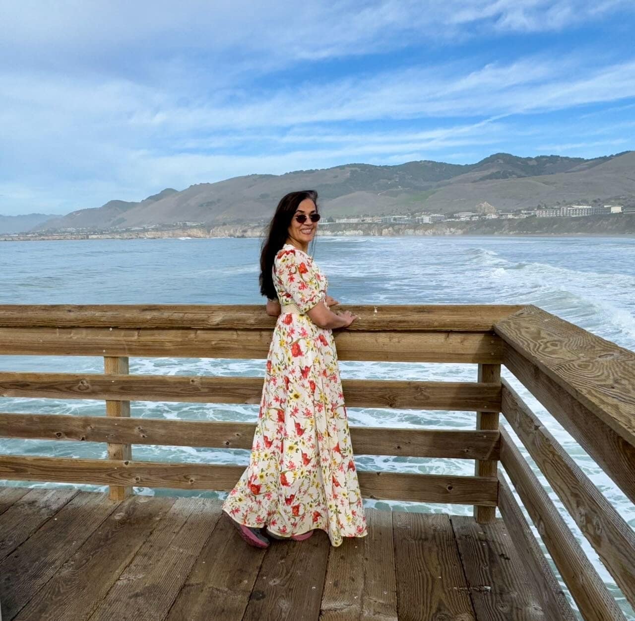 Jyoti in a floral dress standing on the Pismo Beach pier railing looking back over her shoulder with waves and the green coastal hills behind