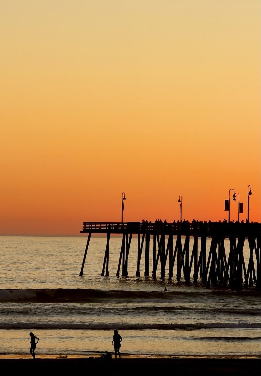 Silhouette of Pismo Beach pier at sunset with the pier structure and lamp posts dark against a deep orange sky and people gathered along the pier