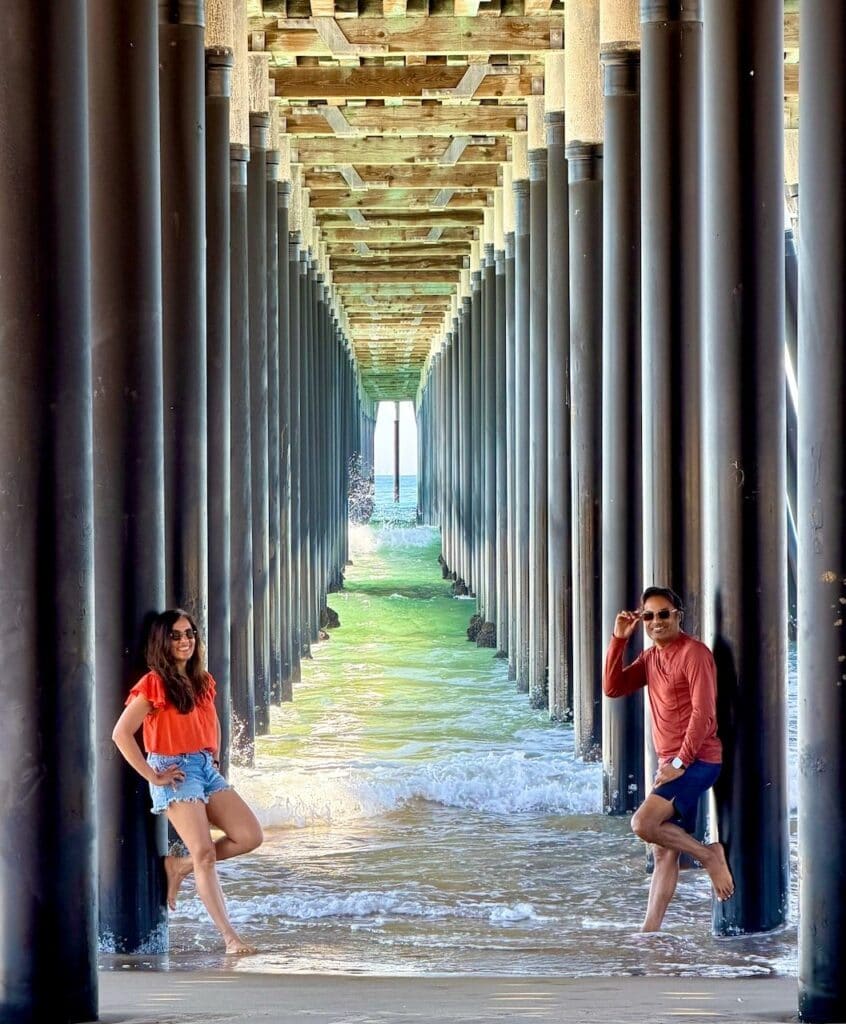 Nirmal and Jyoti jumping with arms raised in the air beneath Pismo Beach pier with waves splashing around them and the ocean visible at the far end