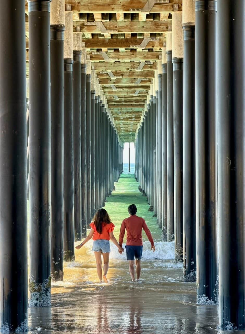 Jyoti and Nirmal leaning against the pillars under Pismo Beach pier with waves washing around their feet and the long perspective of pillars receding behind