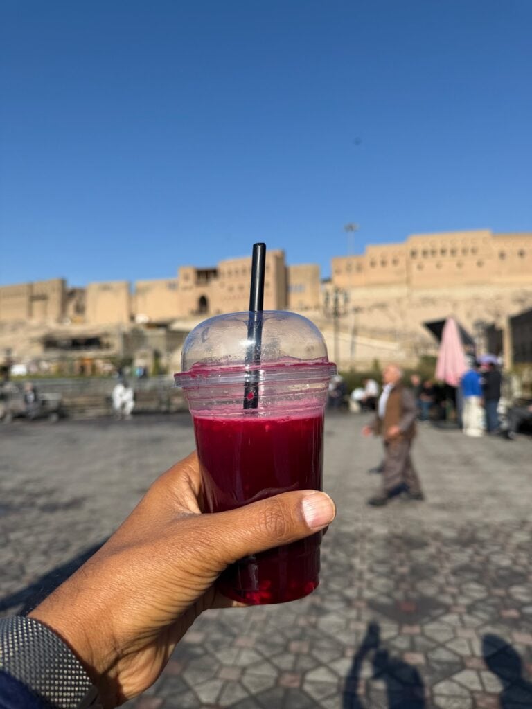 A hand holding a cup of fresh pomegranate juice in front of the ancient Erbil Citadel walls on a sunny day, Iraqi Kurdistan