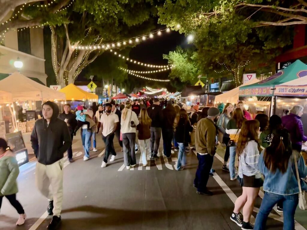Crowded Thursday night farmers market on Higuera Street in downtown San Luis Obispo with string lights in the trees vendor tents and hundreds of people