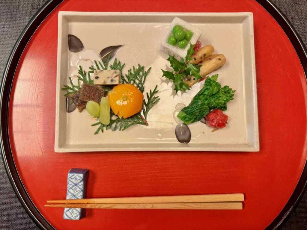 Shojin ryori Buddhist temple cuisine served on a traditional red lacquer tray at Daigo restaurant near Atago Jinja, Tokyo. White ceramic plate with seasonal vegetables, edamame, citrus, and greens, with wooden chopsticks and a blue ceramic chopstick rest.