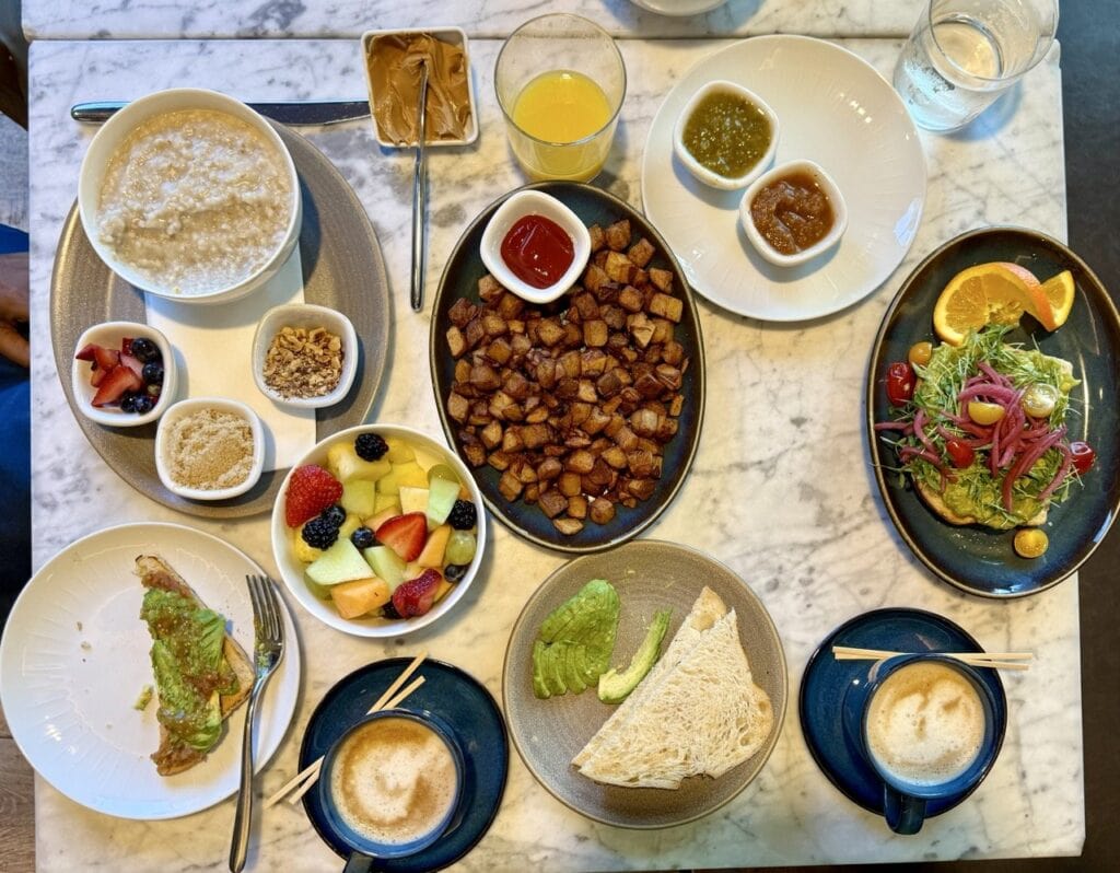 Overhead flat lay of a plant-based breakfast spread at Vespera Resort Pismo Beach including oatmeal avocado toast roasted potatoes fresh fruit lattes and juices on a marble table