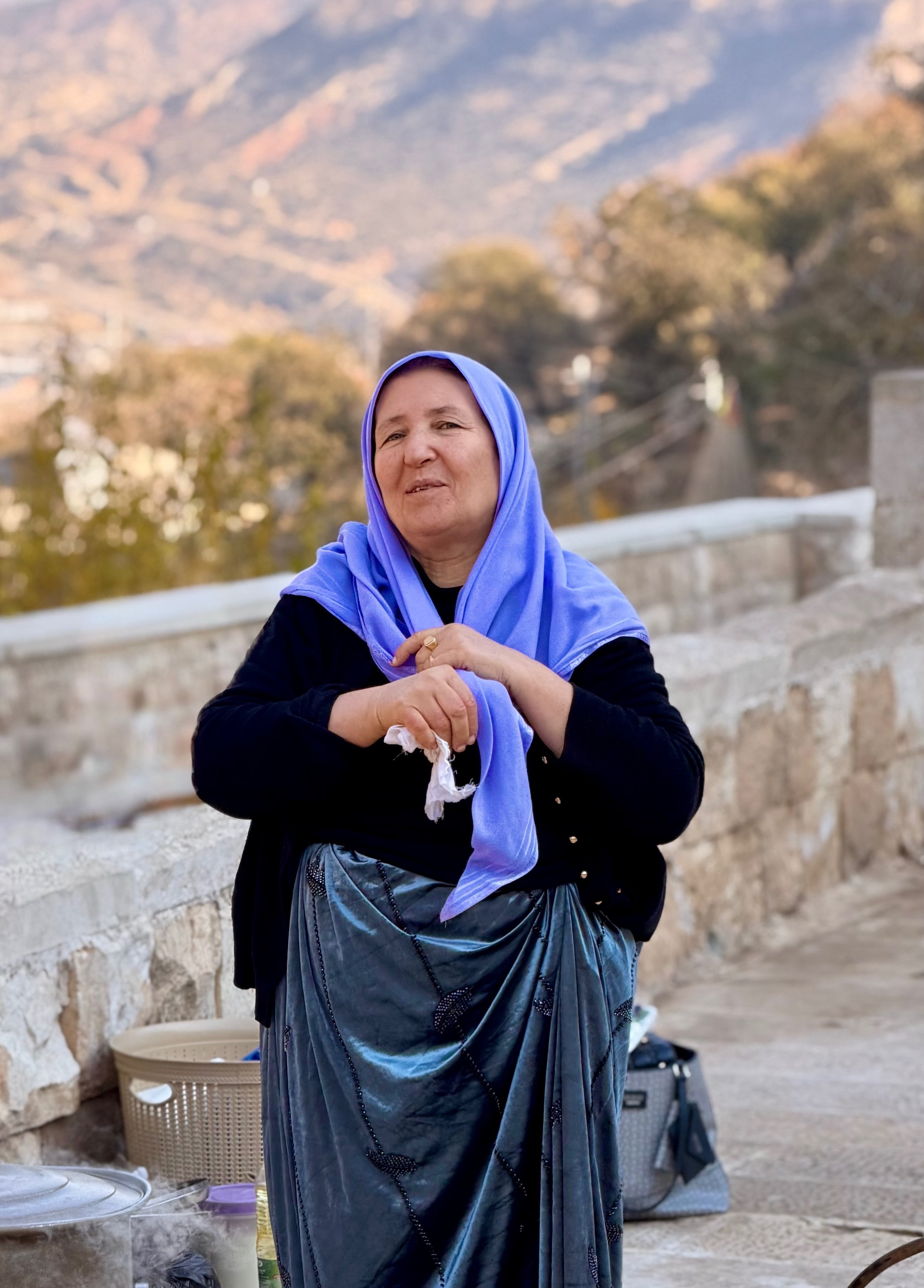 Portrait of a Yazidi woman in a blue headscarf and teal velvet dress smiling at the Lalish temple complex with mountains behind, Iraqi Kurdistan