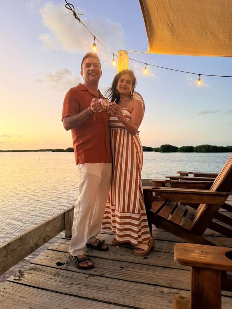Puja and Adi standing on a wooden dock at sunset in San Pedro, Belize, with string lights and calm water in the background