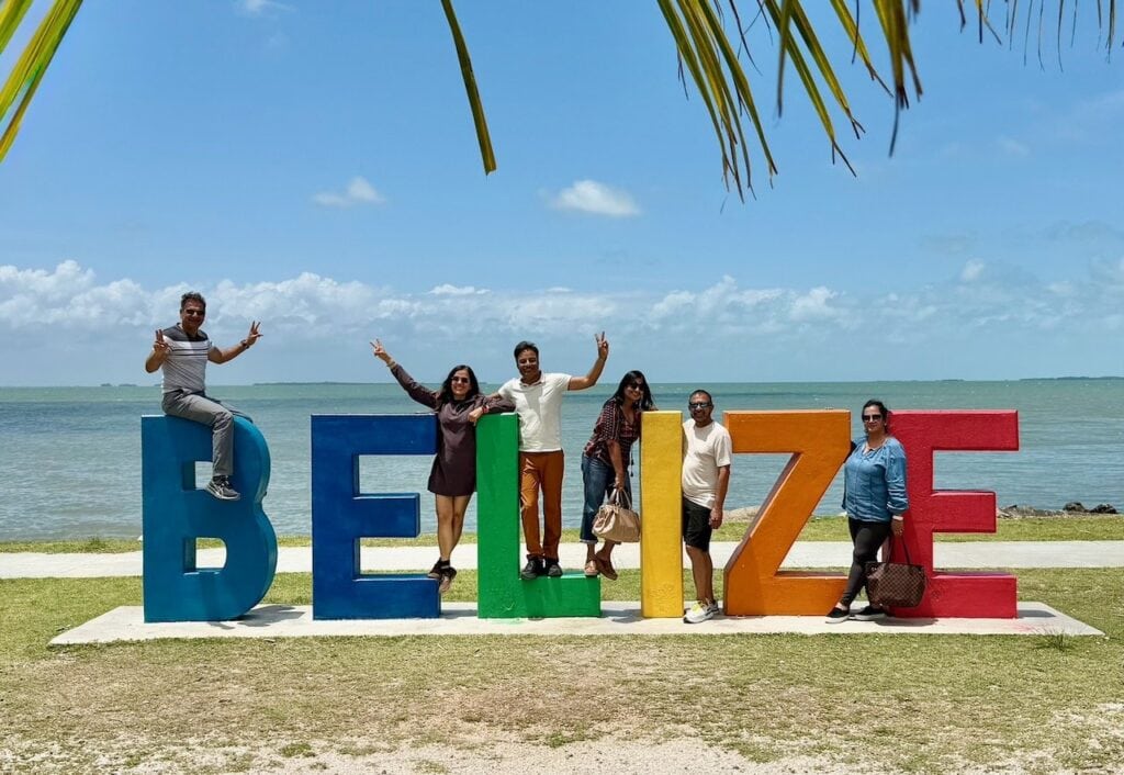 Group of six friends posing at the colorful Belize sign at the Belize City waterfront during a layover