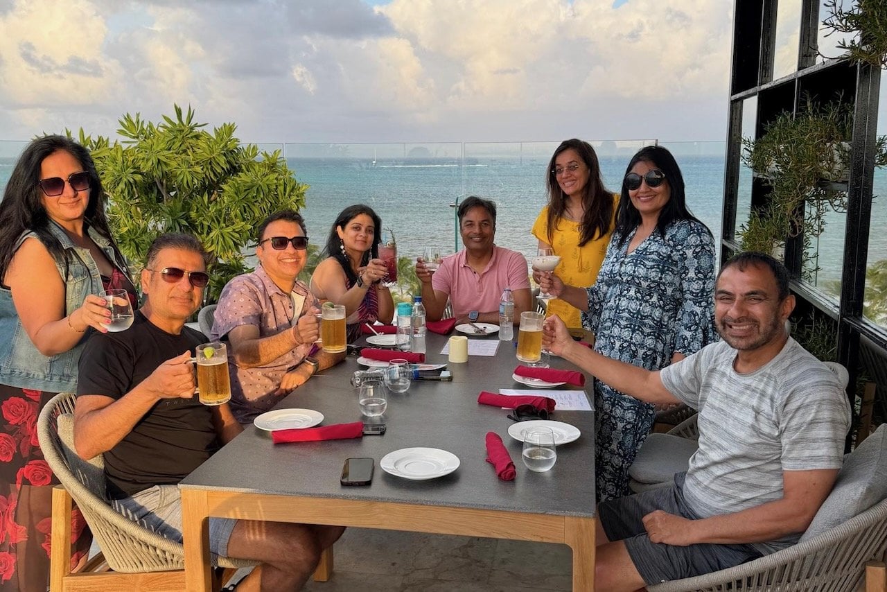 Group of eight friends toasting at dinner at Vista Rooftop Lounge at Alaia Belize, with the Caribbean Sea visible behind them