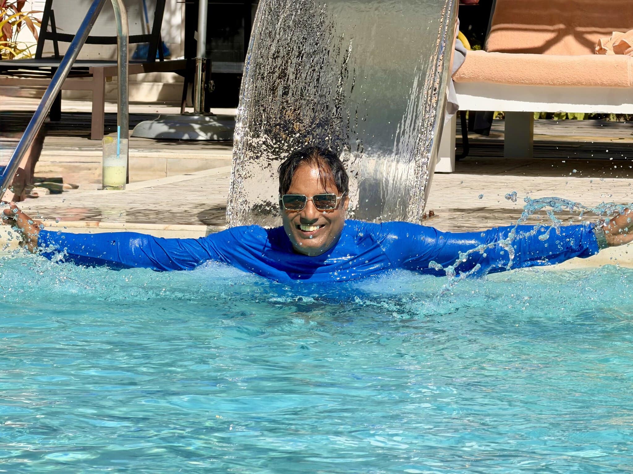 Nirmal standing under a pool waterfall at the Alaia Belize resort with arms spread wide and a big smile