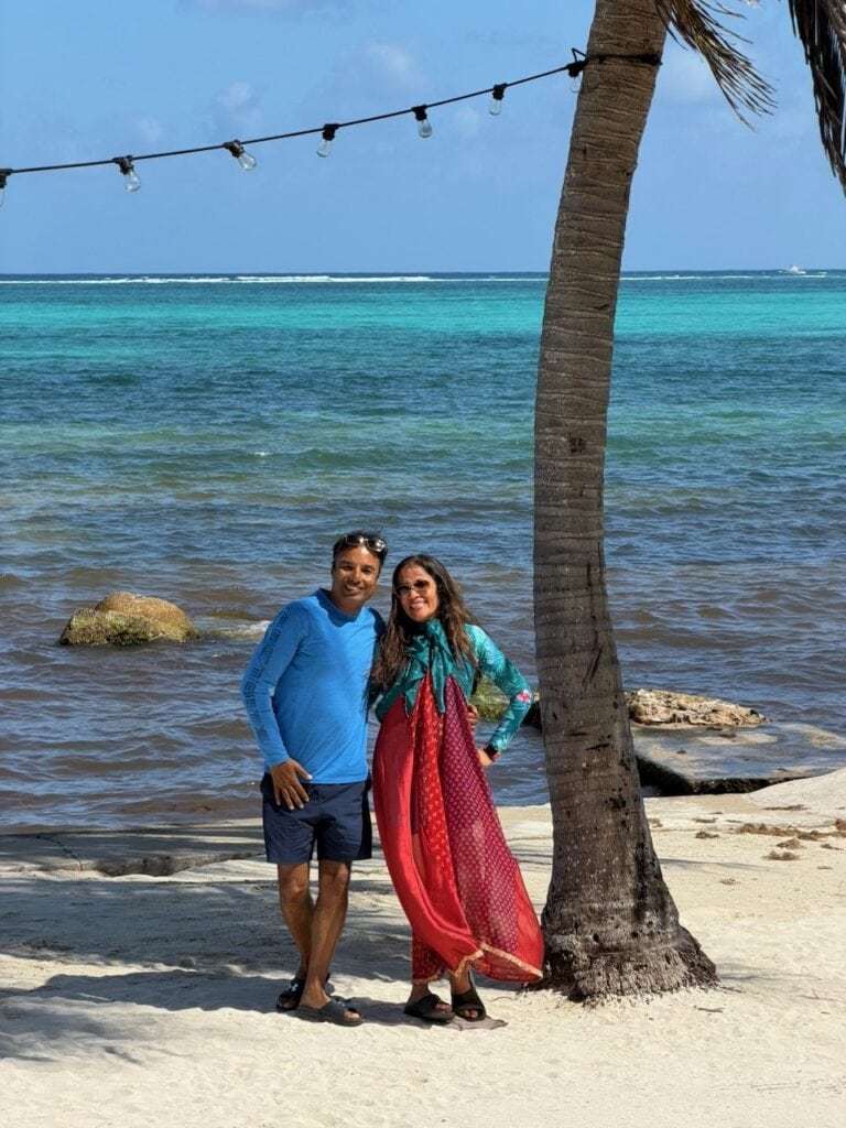 Nirmal and Jyoti standing under a palm tree on the white sand beach at San Pedro, with the turquoise Caribbean Sea behind them