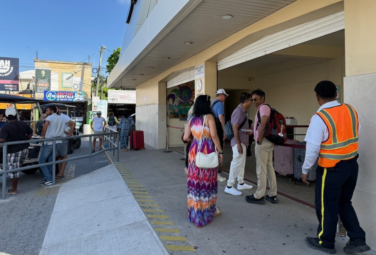 Outside San Pedro Airport terminal with passengers queuing and a golf cart rental shop visible directly across the narrow street