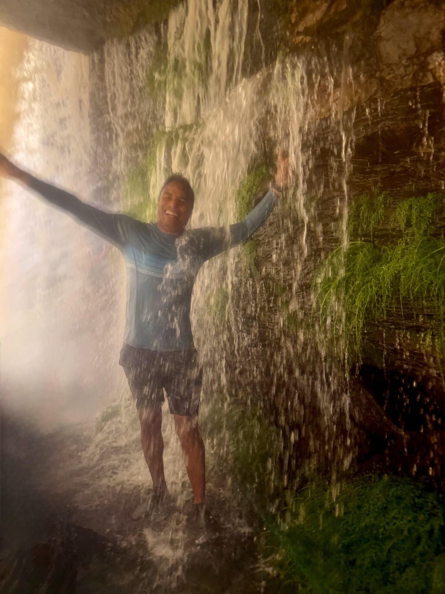 Man standing behind Salto El Sapo waterfall with arms outstretched, soaking wet, smiling