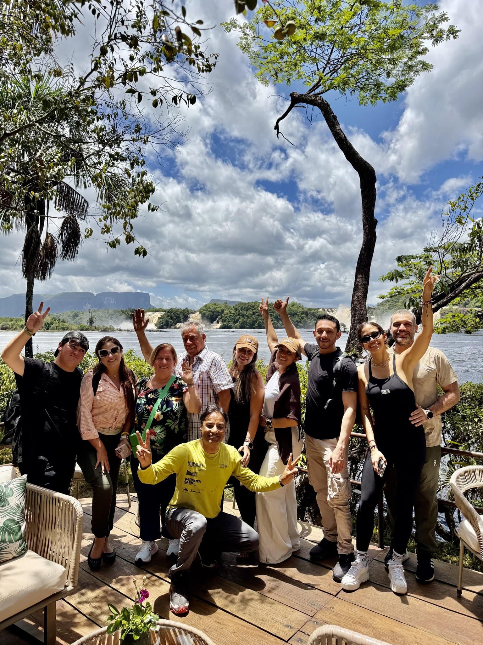 Group of eleven travelers posing on waterfront deck at Canaima with waterfalls and tepui behind them