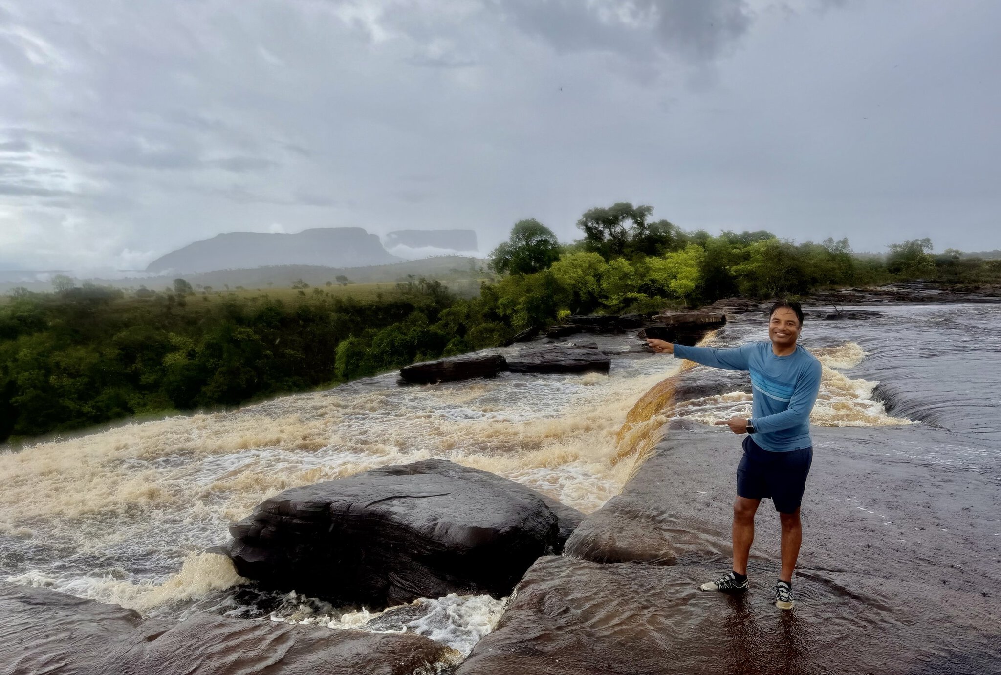 Man pointing and smiling standing on wet rocks above rushing tannin-red river with tepui mesa in background