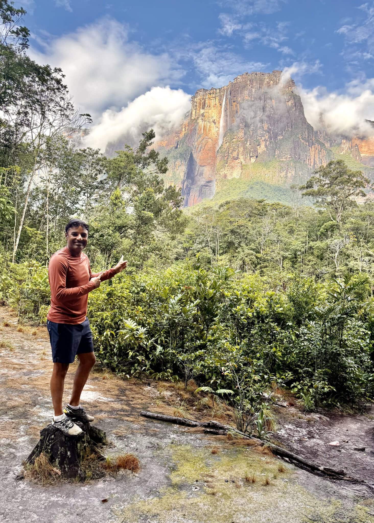 Man giving thumbs up at jungle viewpoint with Angel Falls and cloud-wrapped Auyan Tepui behind him