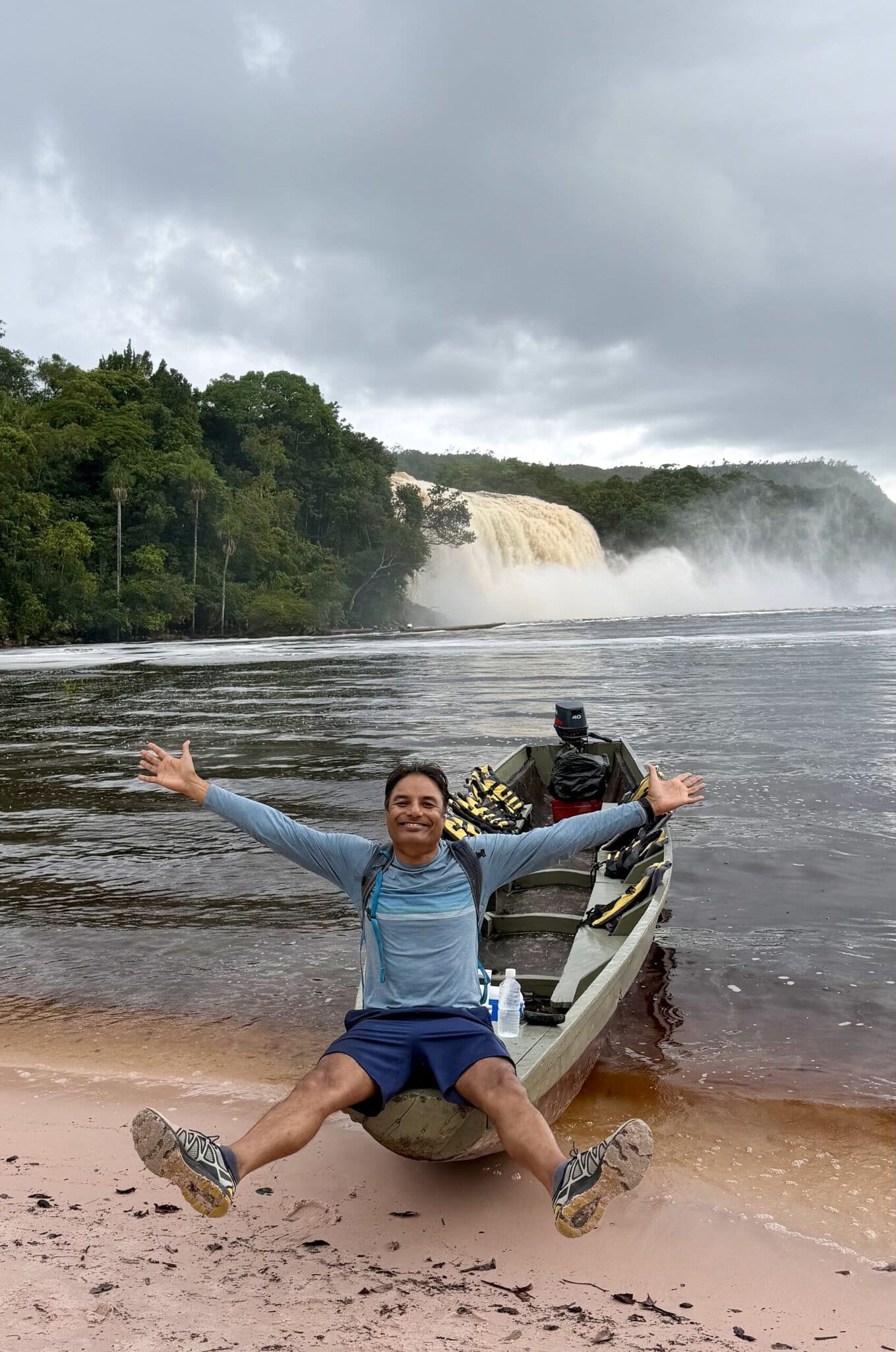 Joyful man sitting arms wide open on bow of motorized canoe with waterfall in misty background