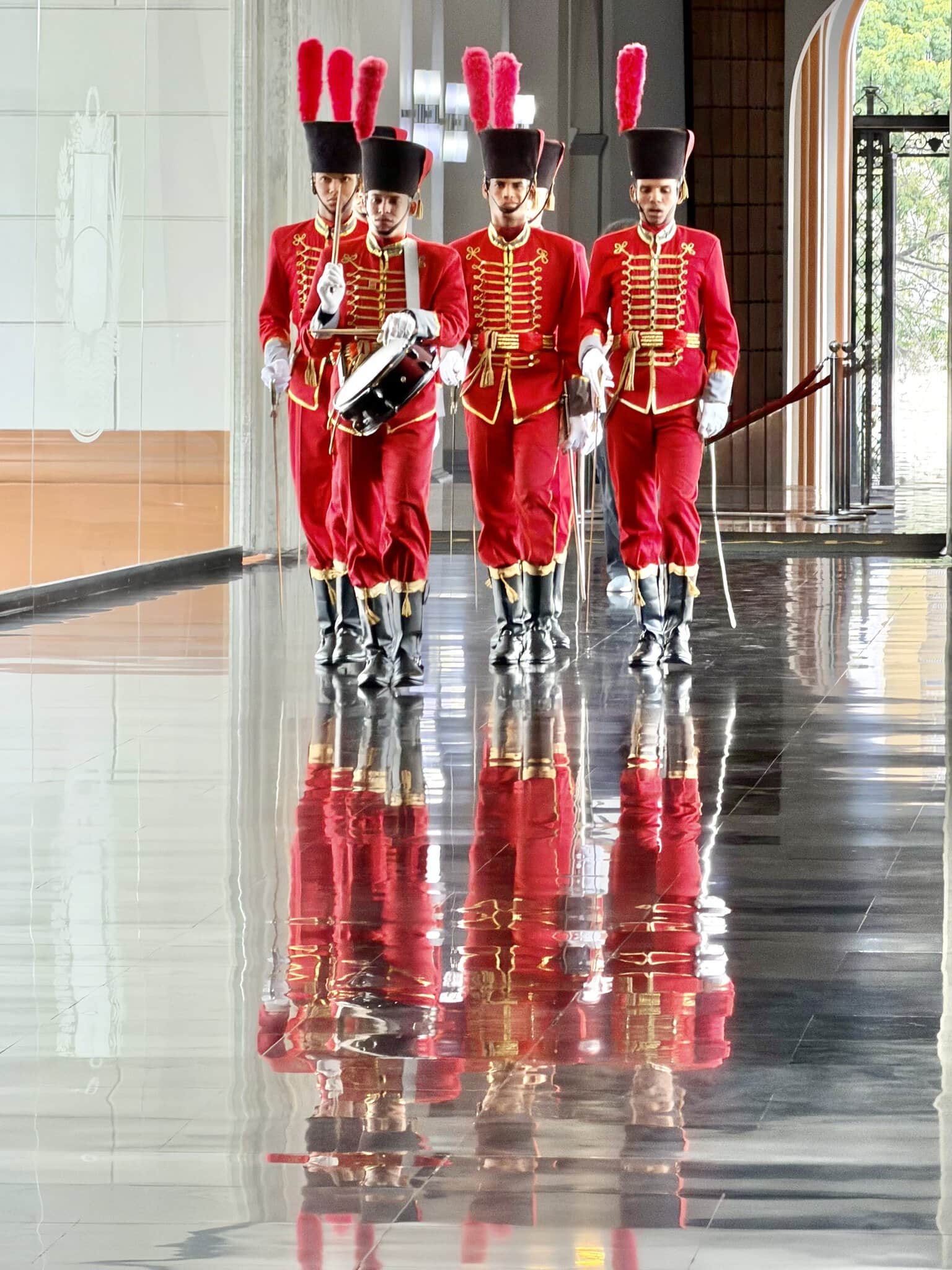 Four guards in red and gold ceremonial uniforms marching on a reflective marble floor in Caracas