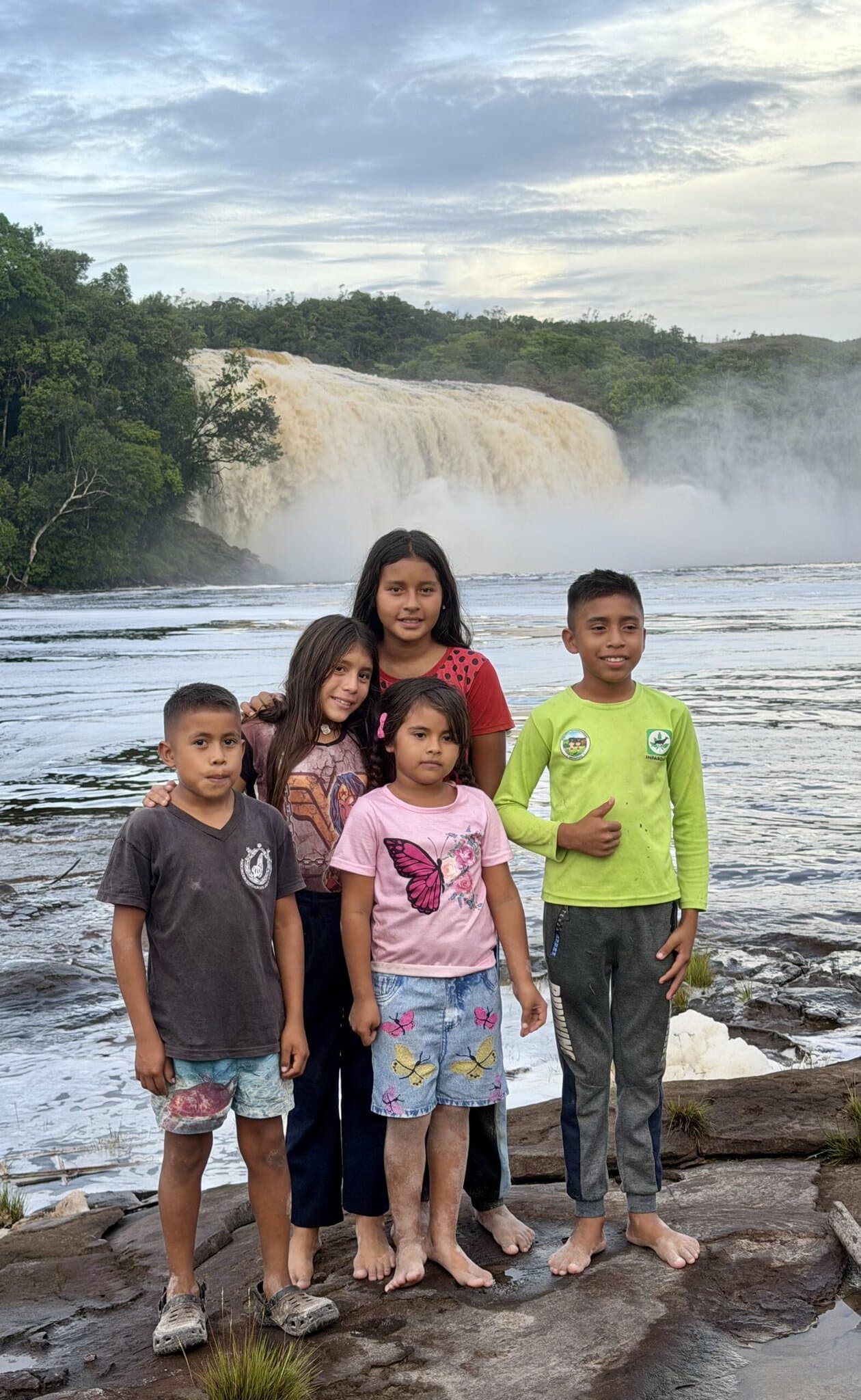 Five Pemon indigenous children smiling together on rocks with massive waterfall and Canaima lagoon behind them