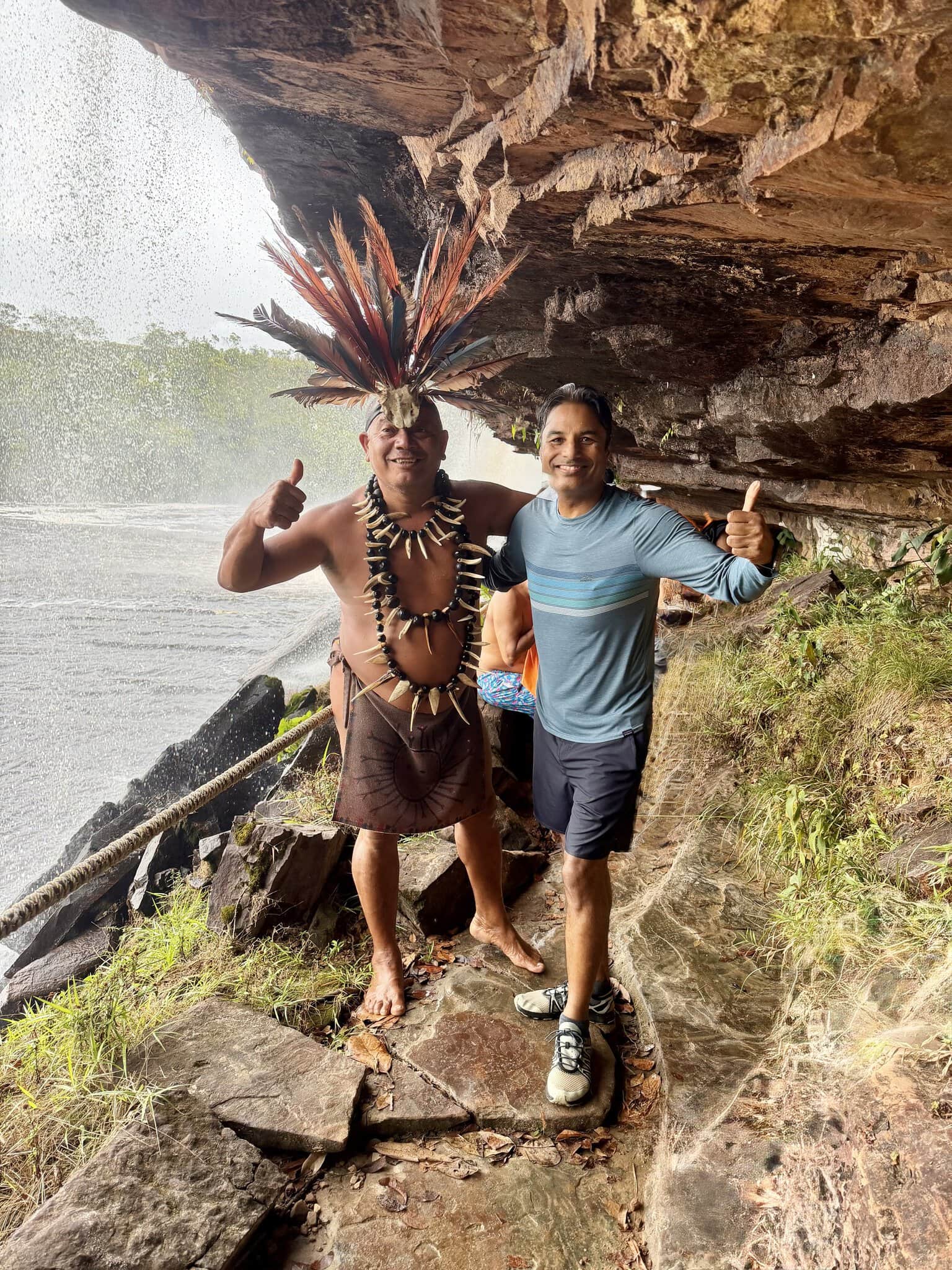 Two men giving thumbs up beside Angel Falls waterfall, one in traditional Pemon indigenous dress