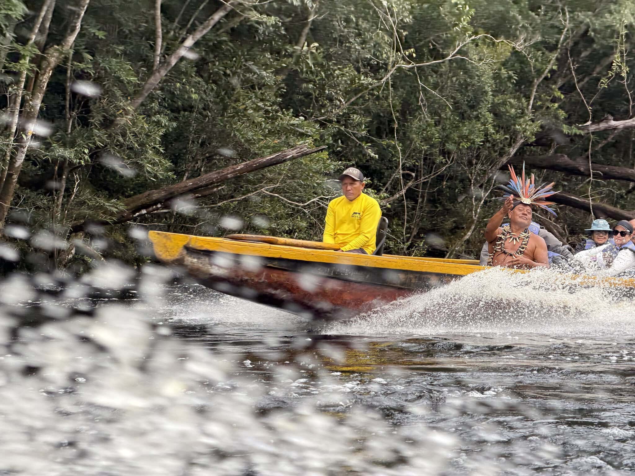 Pemon indigenous guide wearing traditional feather headdress and beaded necklace on a yellow motorized canoe in Canaima