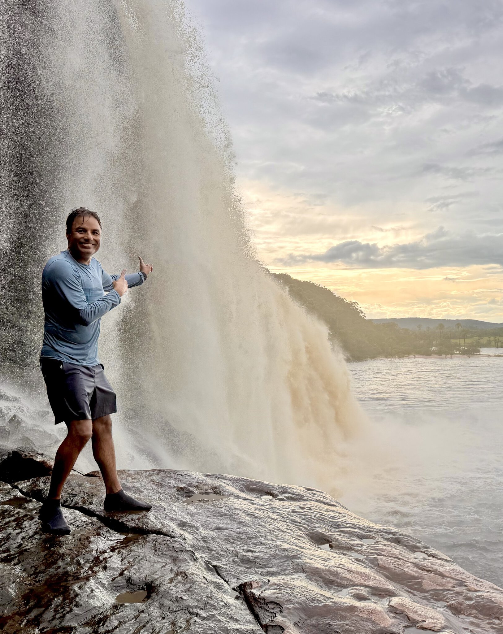 Man pointing thumbs up standing on wet rocks beside massive curtain of Salto el Sapo waterfall Venezuela at sunset