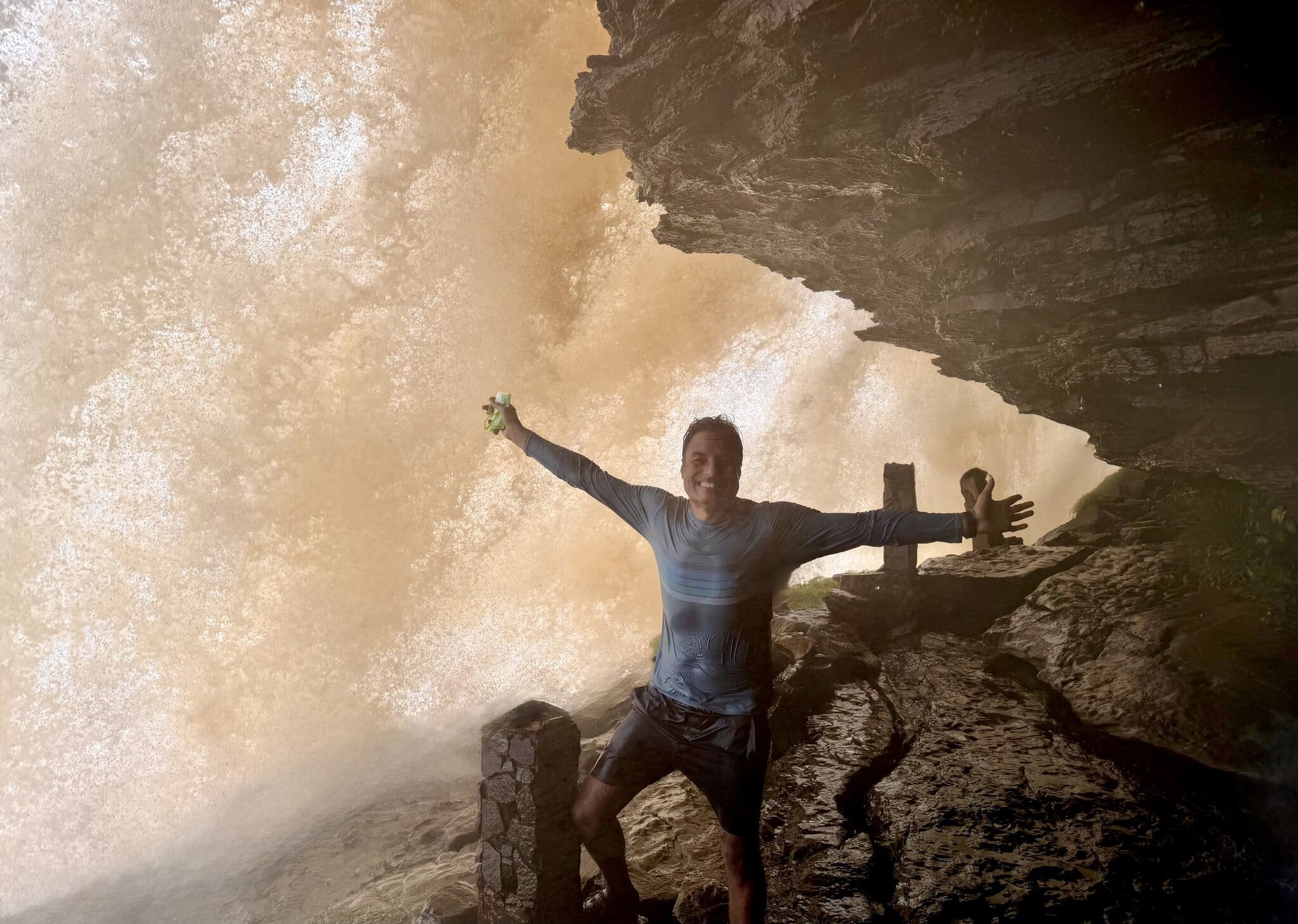 Man with arms spread wide standing behind the thundering curtain of Salto el Sapo waterfall in Canaima