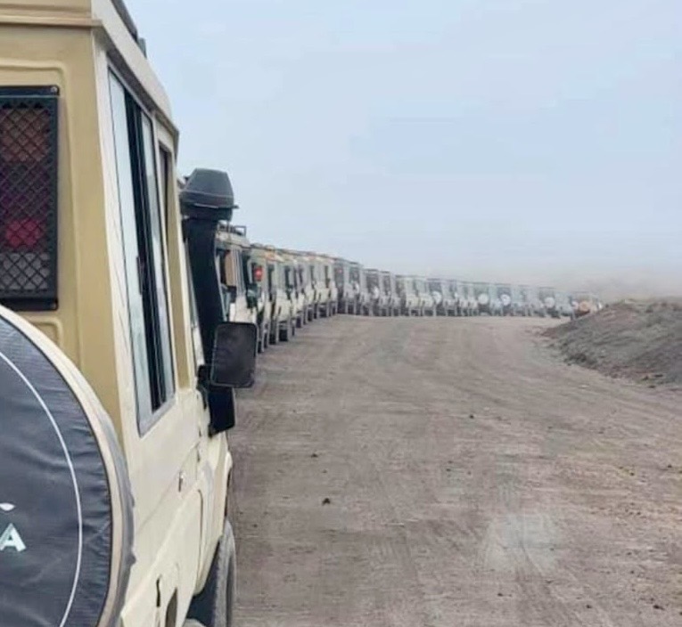 A long line of branded safari vehicles descends the dusty track into Ngorongoro Crater, stretching into the distance